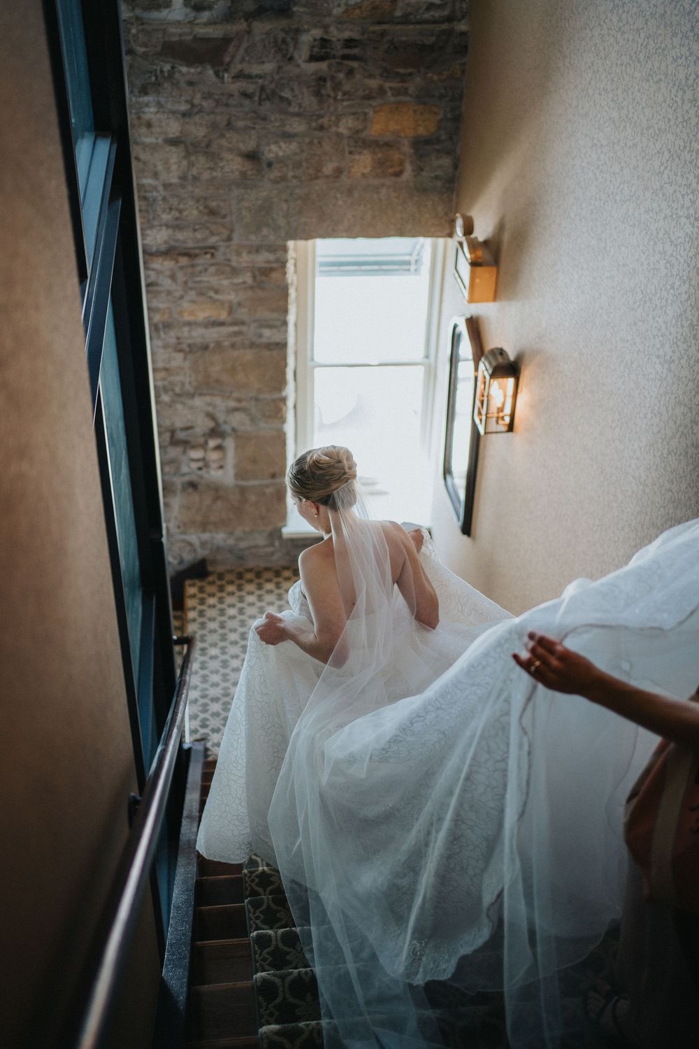A flowing white wedding dress being adjusted in a rustic room with stone walls and natural light.