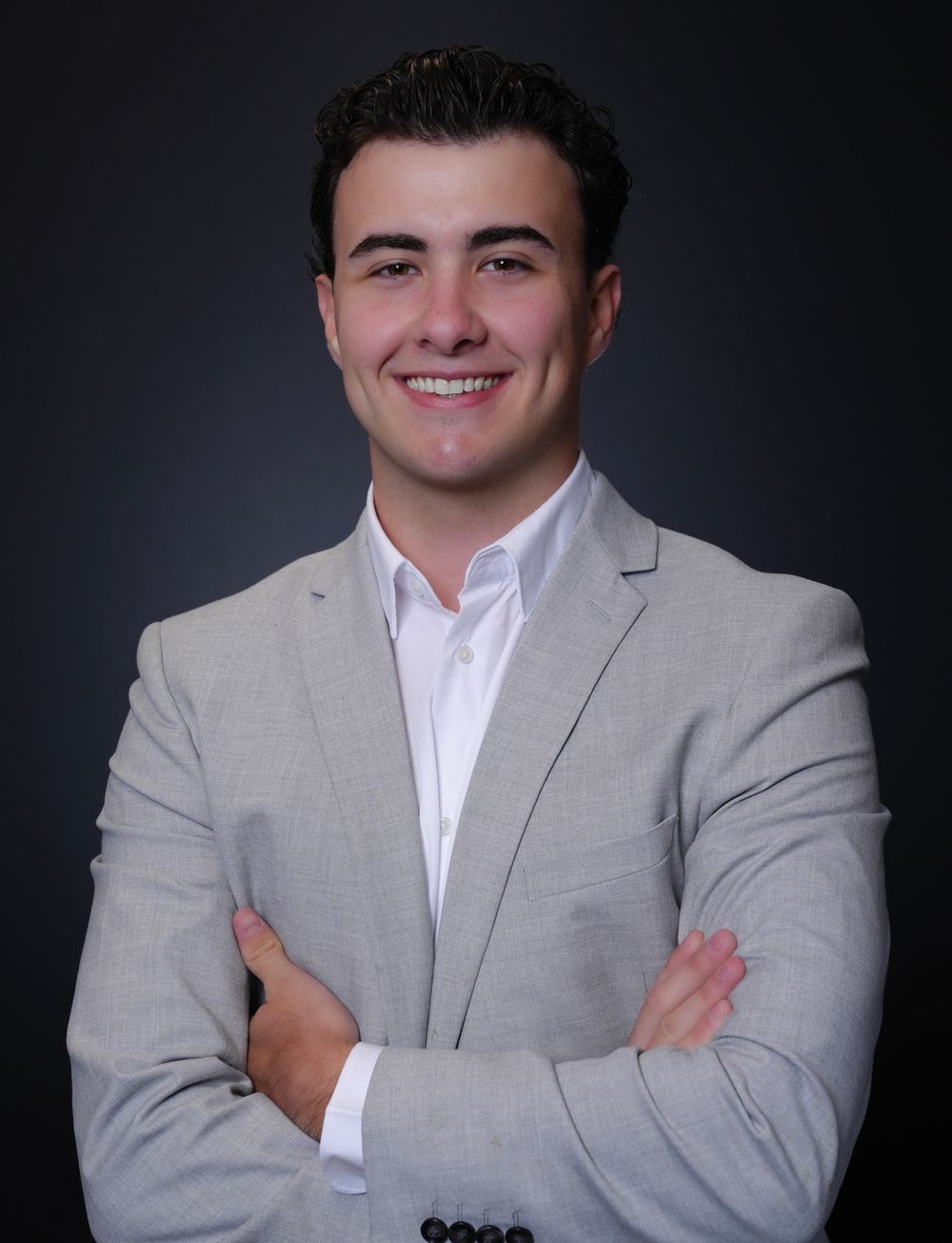 Young professional man in gray suit smiling with arms crossed against dark background.