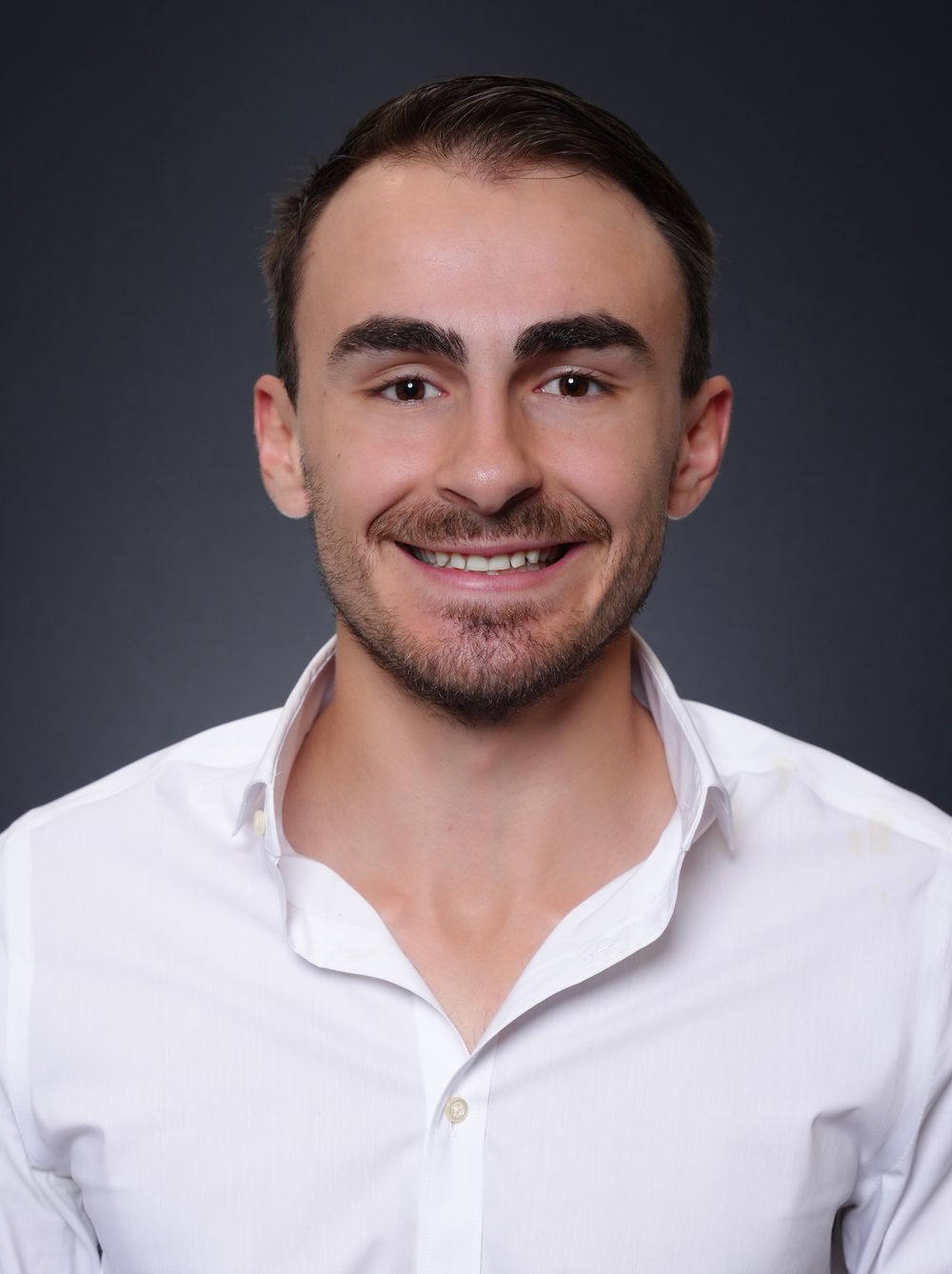 Young professional man smiling in white shirt against dark background, headshot portrait photo.