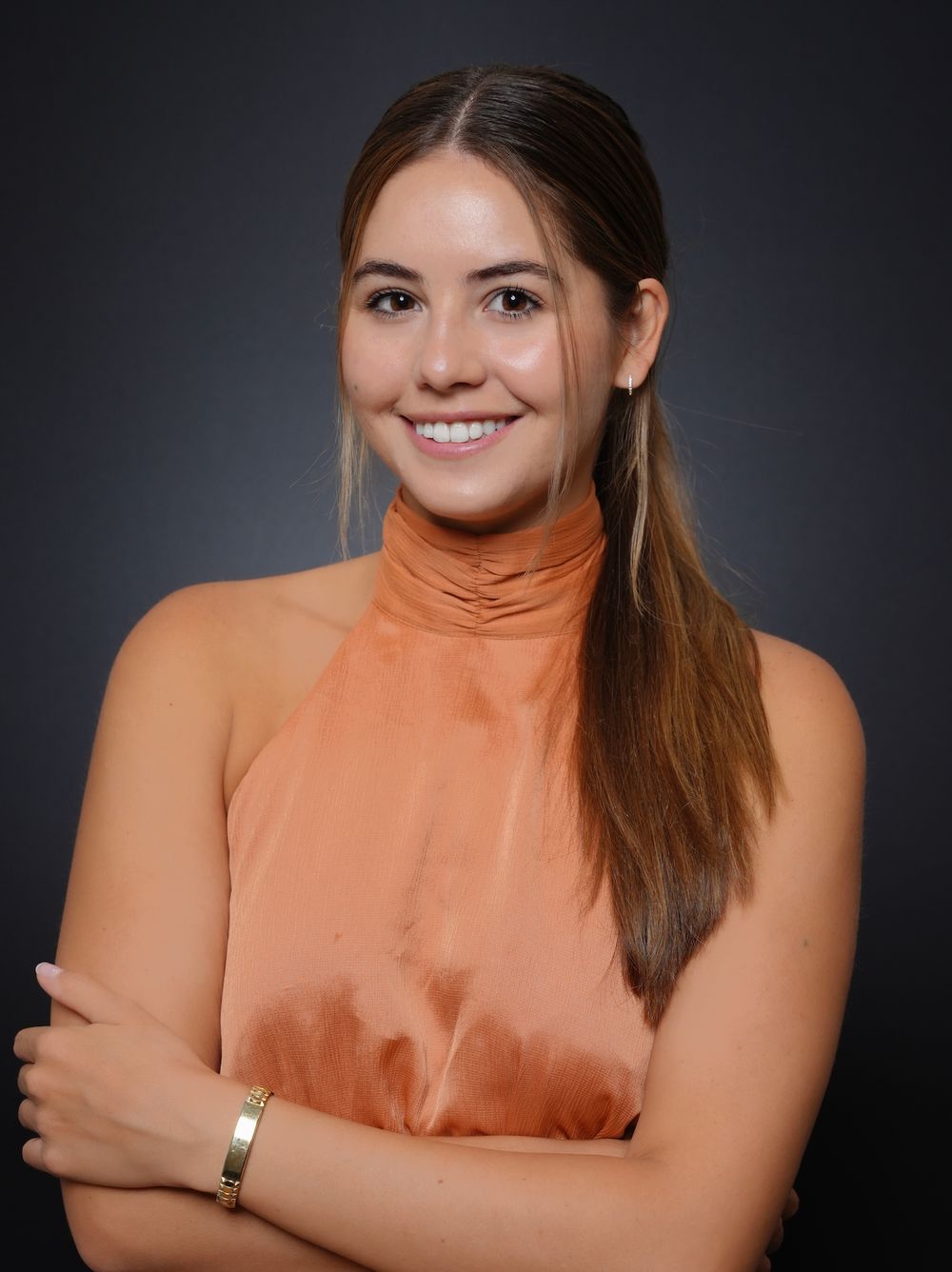 Young woman in peach satin turtleneck top smiling with arms crossed, wearing gold bracelet against dark background.