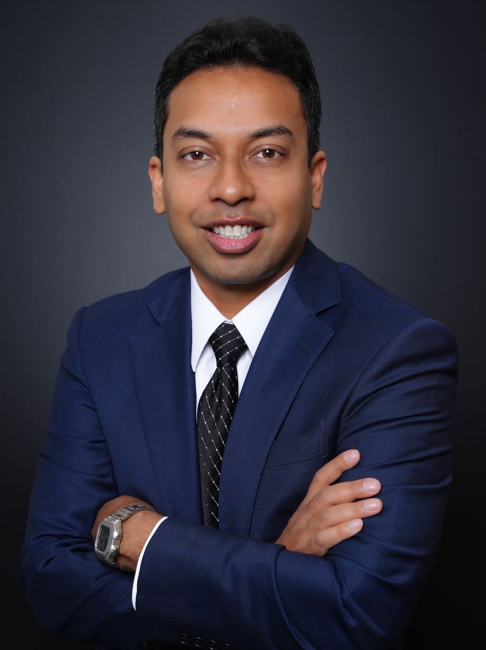 Professional headshot of a smiling man in a navy blue suit and tie with arms crossed against a dark background.