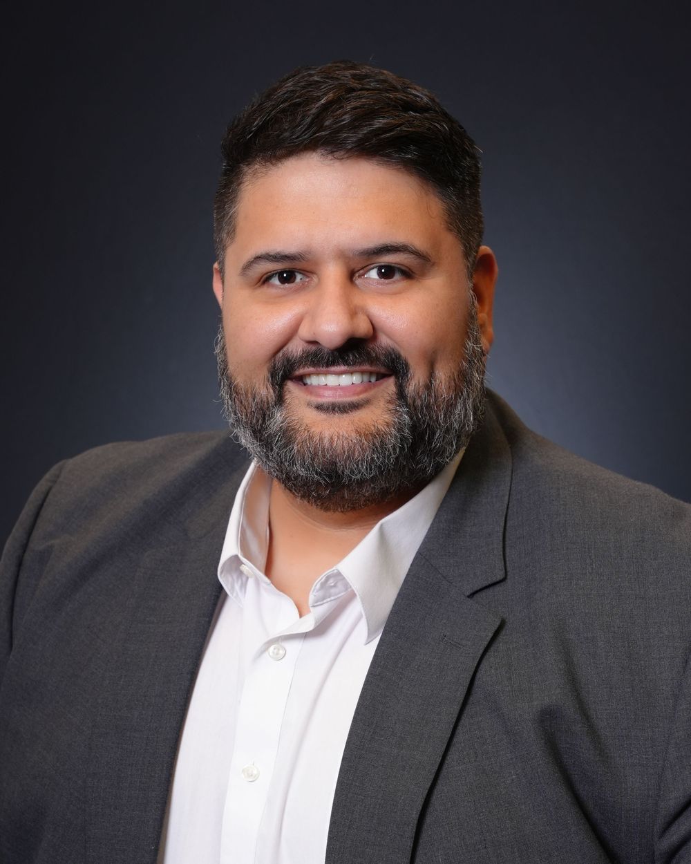 Professional headshot of person in white shirt and dark suit jacket against dark gray studio background.