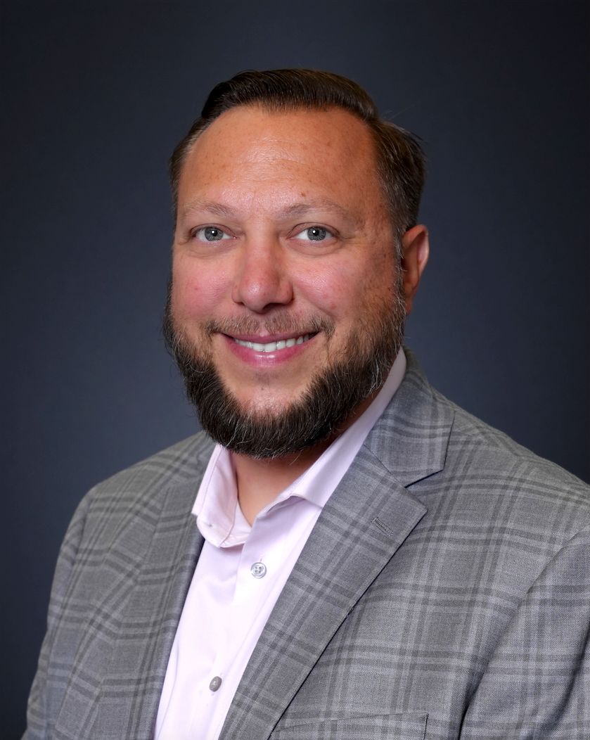 Professional headshot of a person in a gray plaid blazer and white shirt smiling warmly against a dark background.