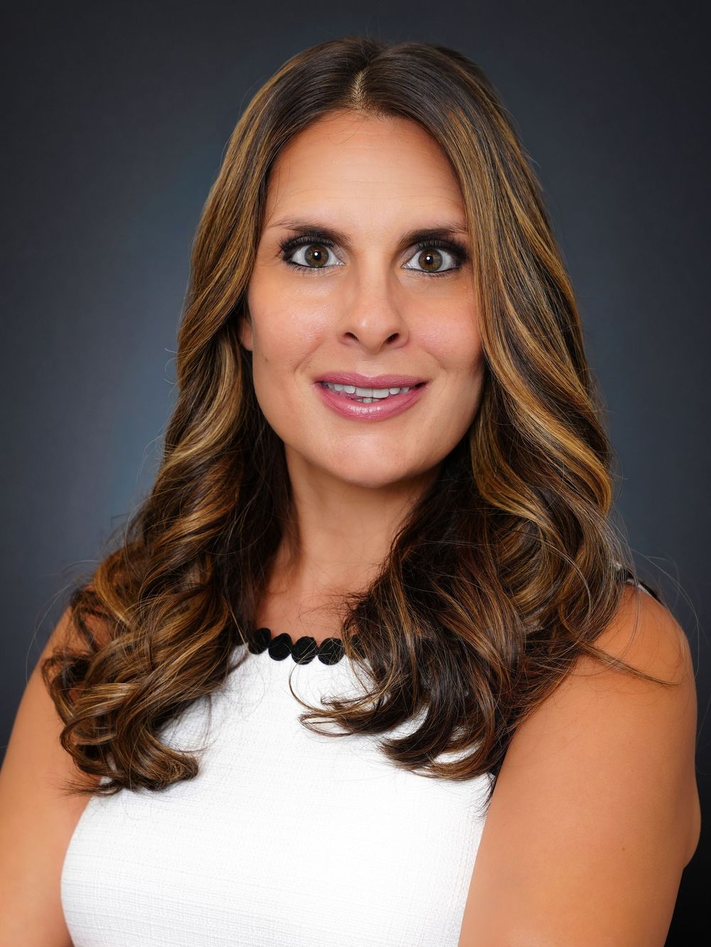 Professional headshot of a smiling woman with long brunette highlighted hair wearing a white top and black beaded necklace.
