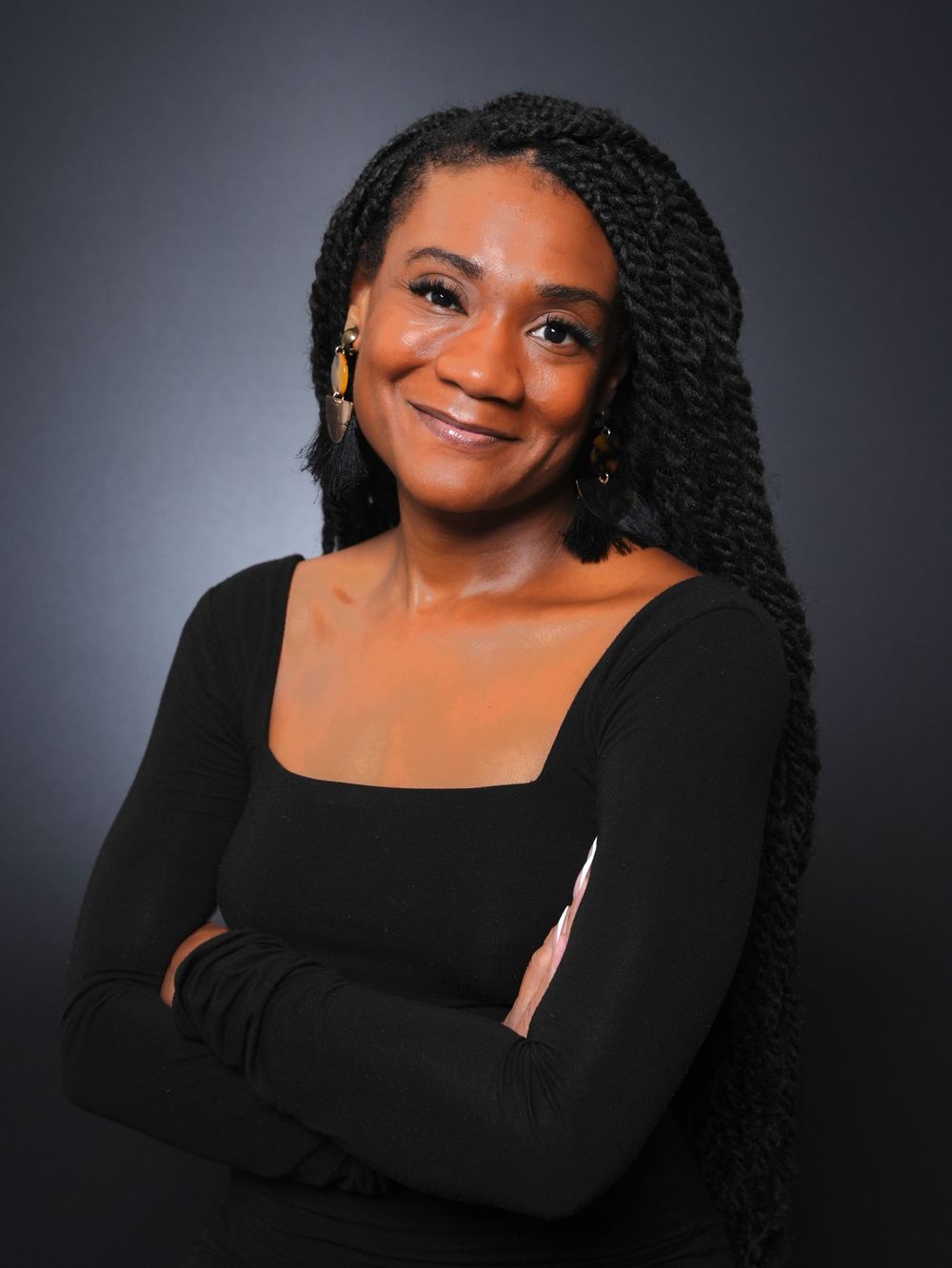 A smiling woman with long braids wearing a black top, arms crossed, posing confidently against a dark gray background.
