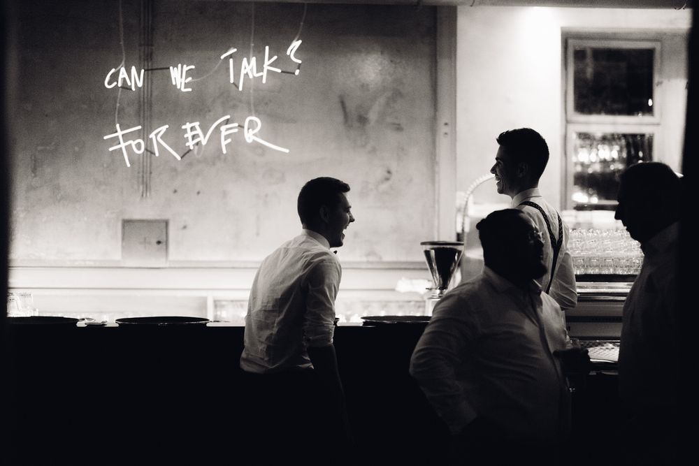 Gäste lachen an der Bar während der Hochzeitsparty, Beleuchtung „Can we talk forever?“ im Hintergrund.