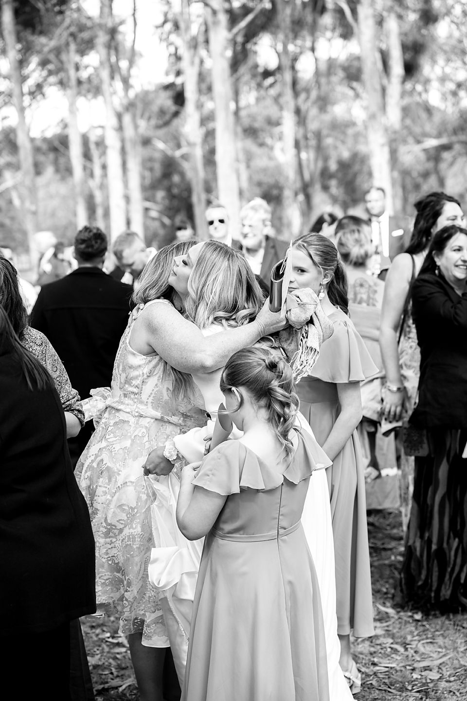 Emotional black and white photo of wedding guests gathered outdoors in a wooded setting during a ceremony.