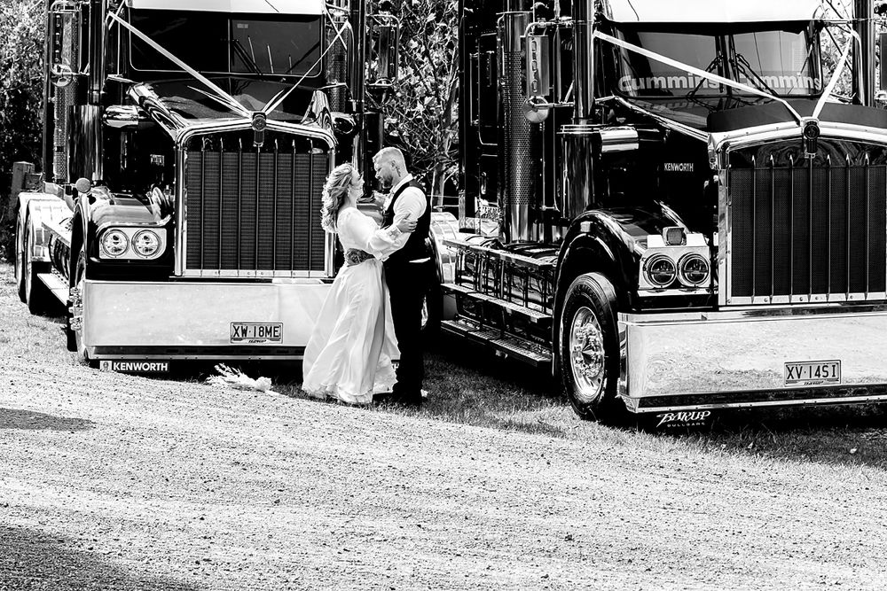 A black and white photo shows a bride and groom walking beside a vintage car at a rustic outdoor wedding.