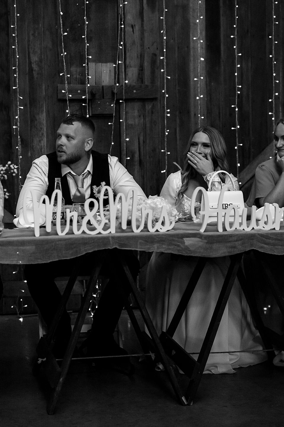 A black and white photo of people seated at a long table with string lights visible in the background.