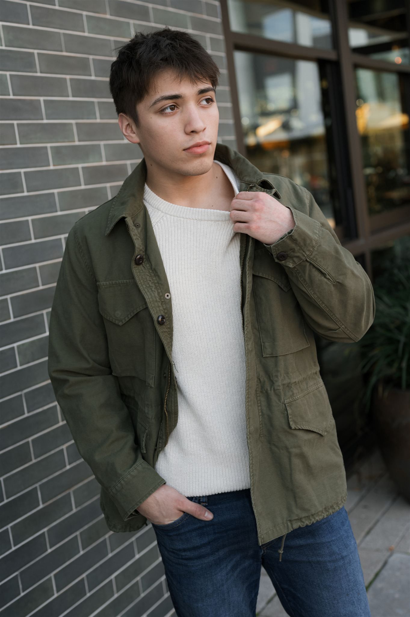 Person in olive green utility jacket and white sweater poses casually against brick wall outdoors.