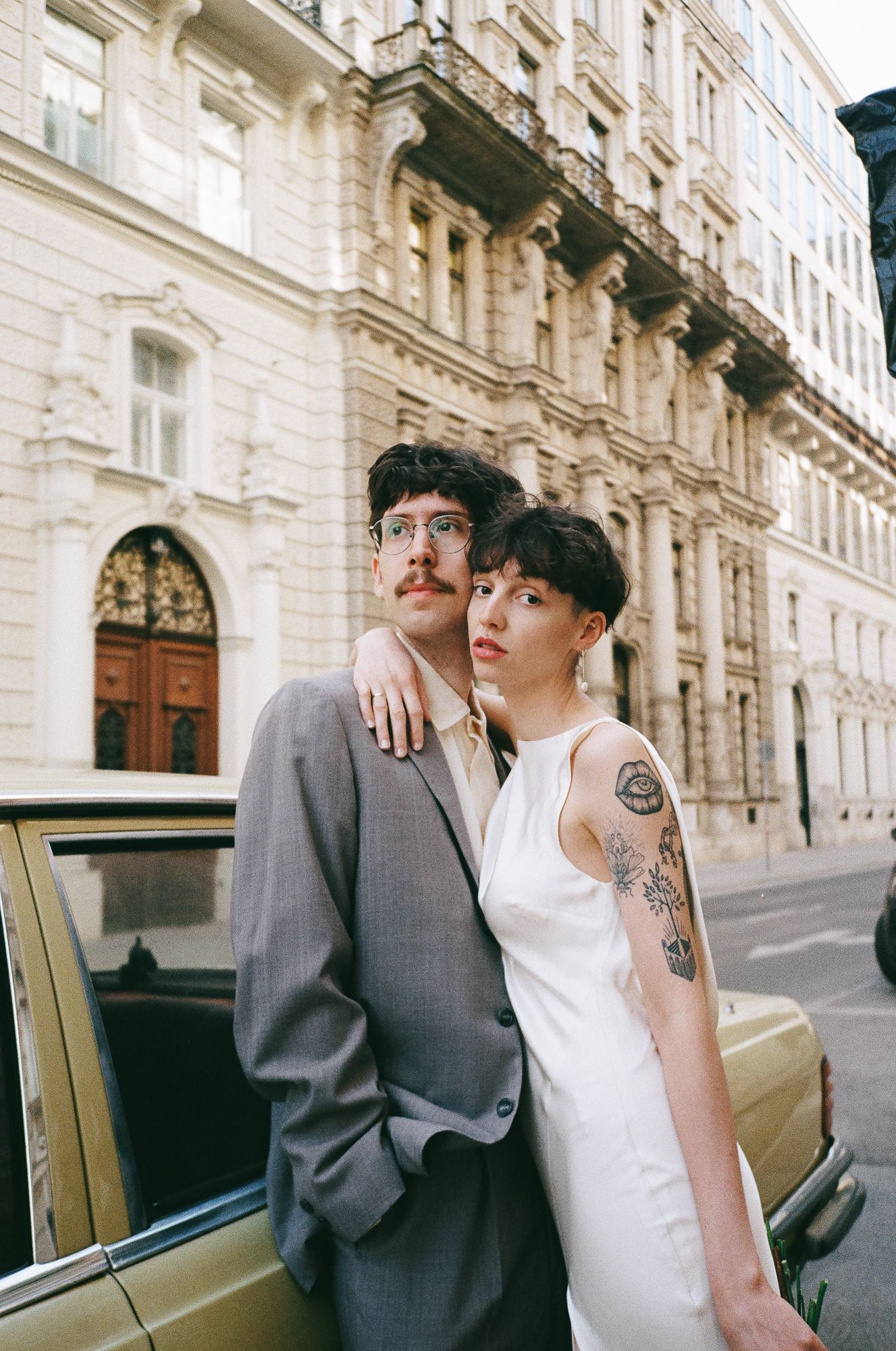 A couple poses together near a vintage car on a European street with classic architecture in the background.