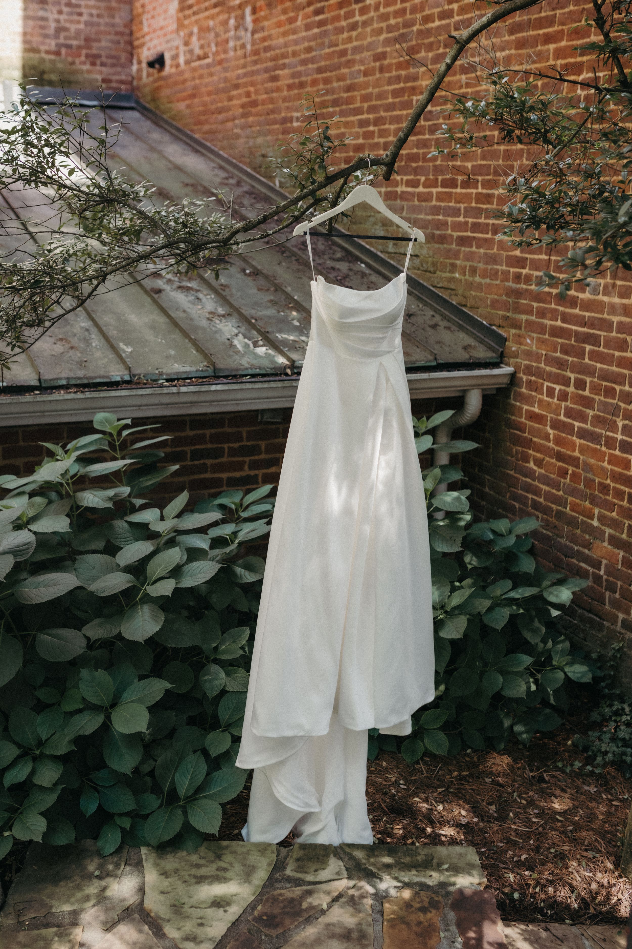 Elegant white wedding dress hanging on a branch against a rustic brick wall with green foliage.