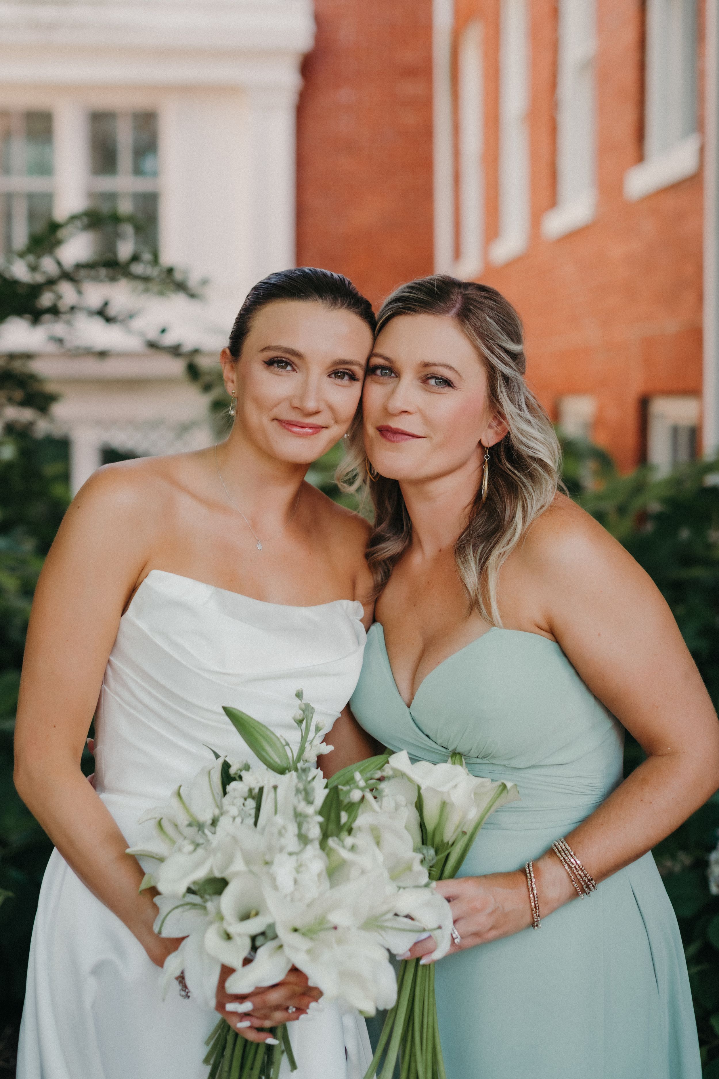 Two women in formal dresses pose together with a white floral bouquet against a brick building.