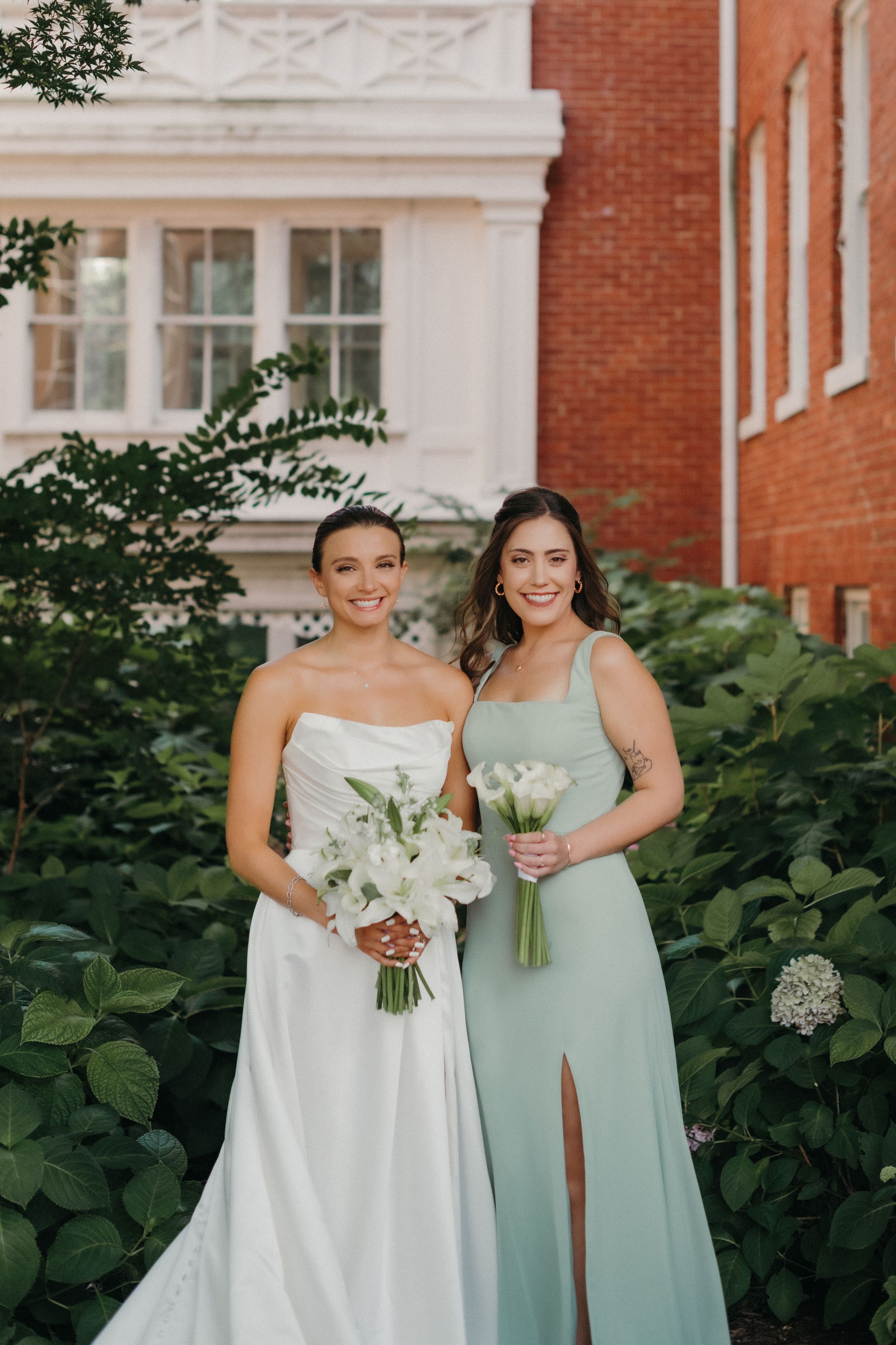 Bridesmaids in white and sage green dresses pose together outside a brick building with white trim.