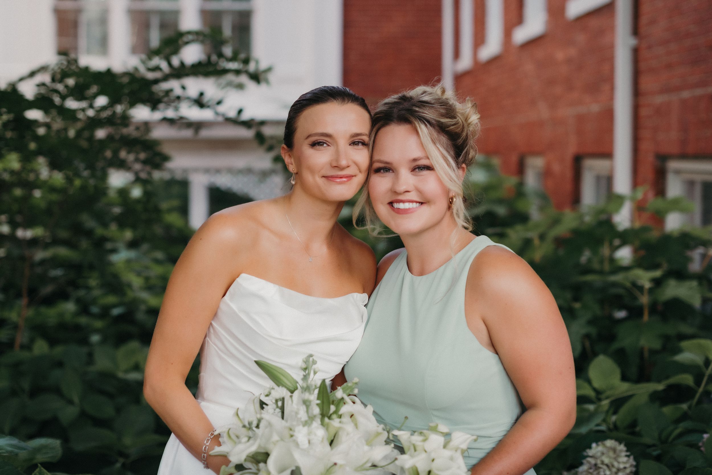 Two women smile together at a wedding, with one in a white bridal gown holding white flowers and one in mint green.