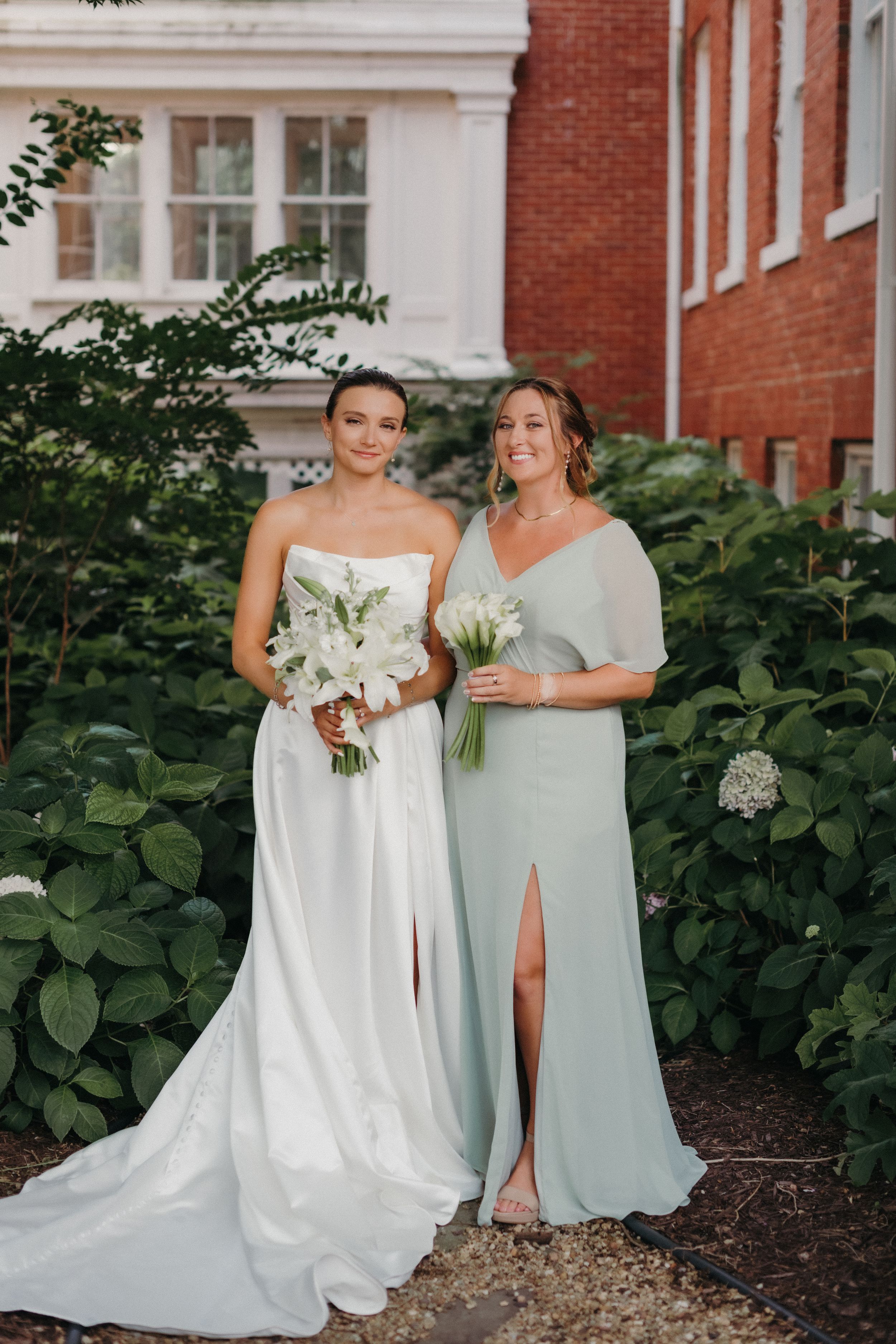 Wedding party members in elegant white and sage green dresses pose together against a brick building backdrop.