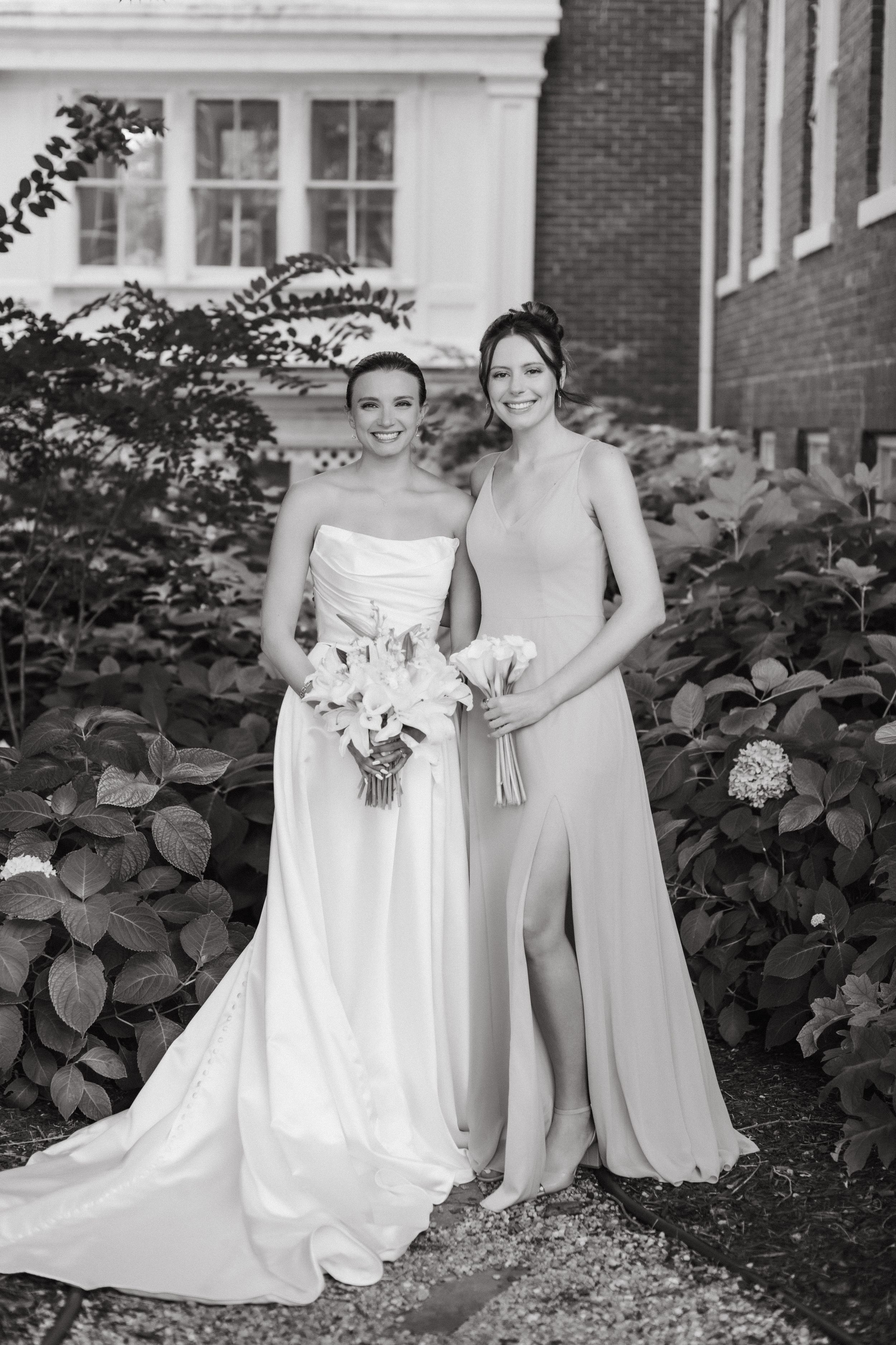Bridesmaids in elegant white and mint green dresses pose together against a brick building backdrop.