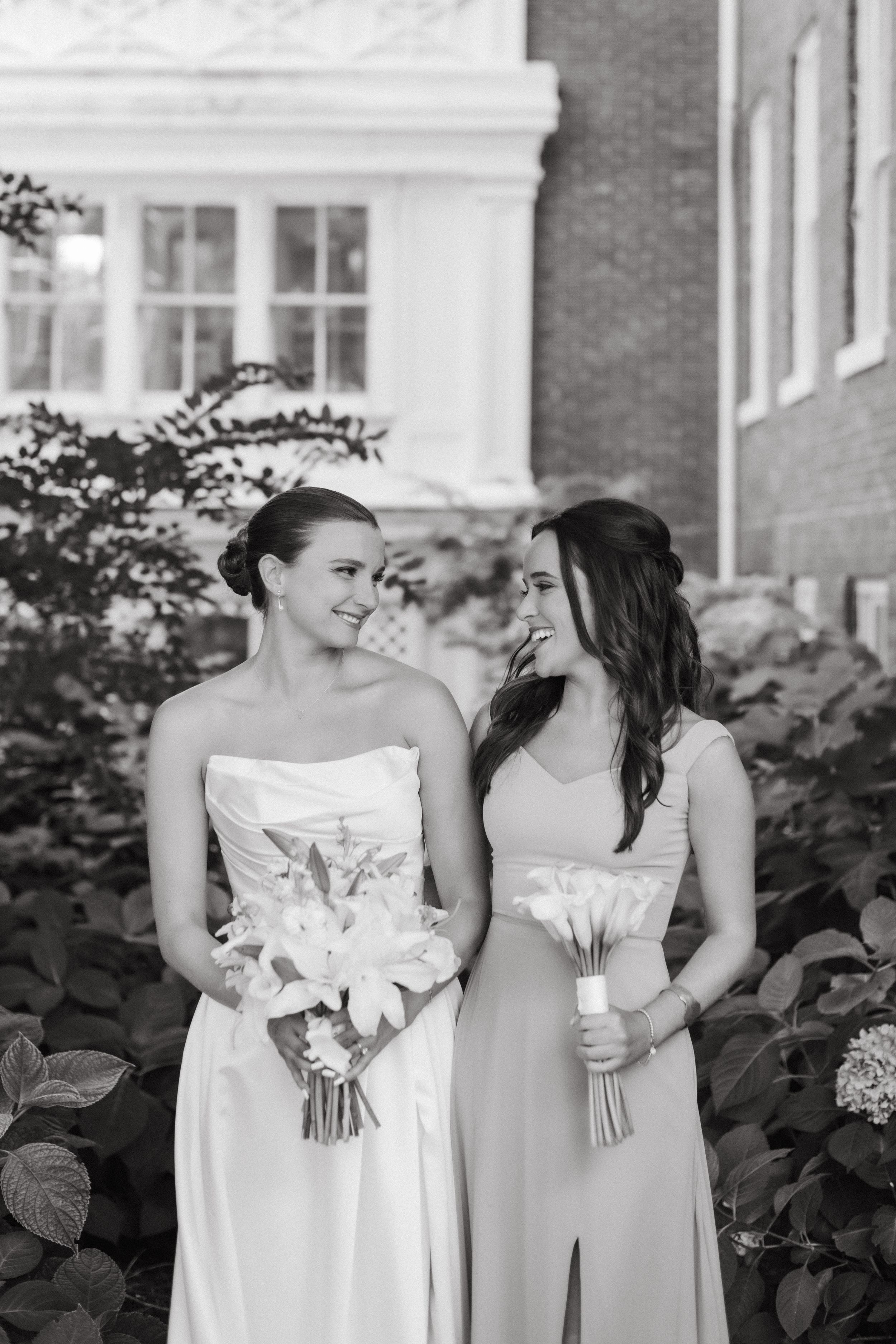 Black and white photo of bridesmaids sharing joyful moments in white and sage gowns by a brick wall.