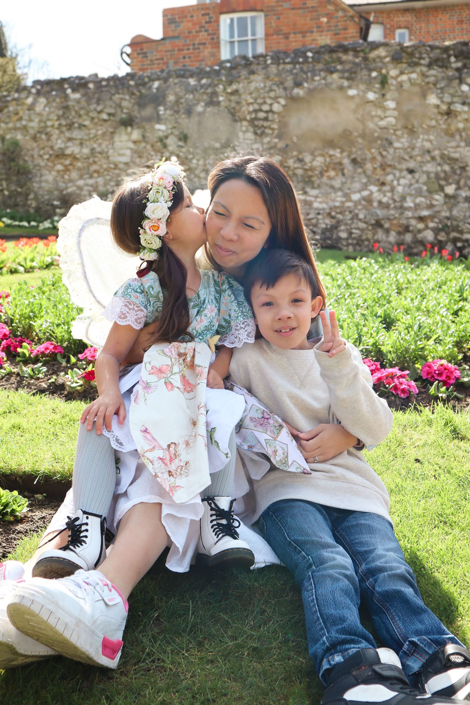 Family enjoying a sunny day together on grass near a brick wall with blooming flowers.