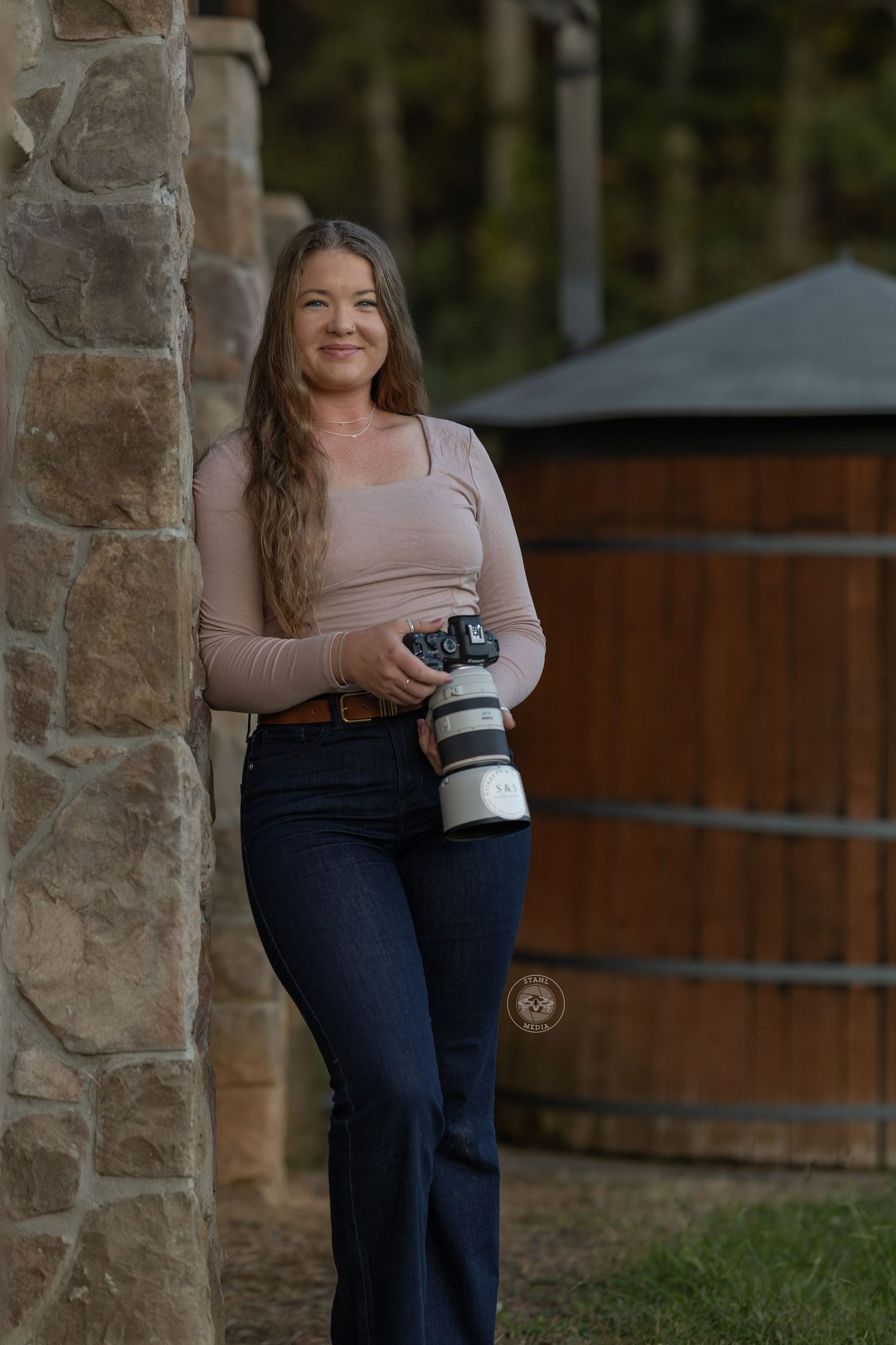 Professional photographer in jeans and pink top holds camera with telephoto lens outdoors by stone pillars.