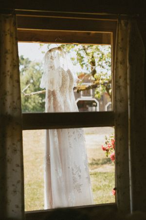White wedding dress hanging in window frame of rustic cabin with natural light.