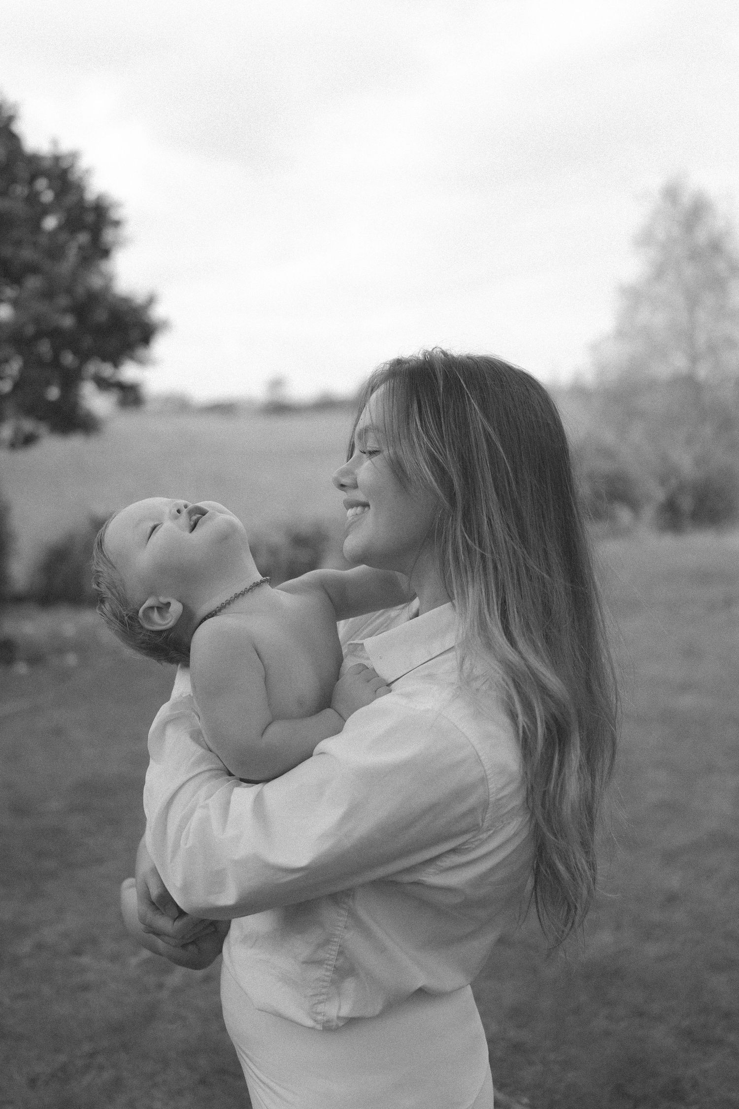 Black and white photo of smiling mother holding laughing baby outdoors in green field in New Zealand.