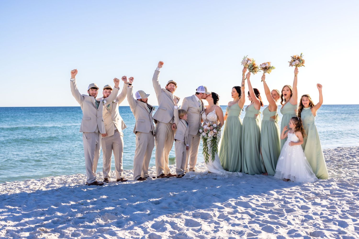 A wedding party in light colored attire poses together on a white sandy beach with ocean waves in the background.