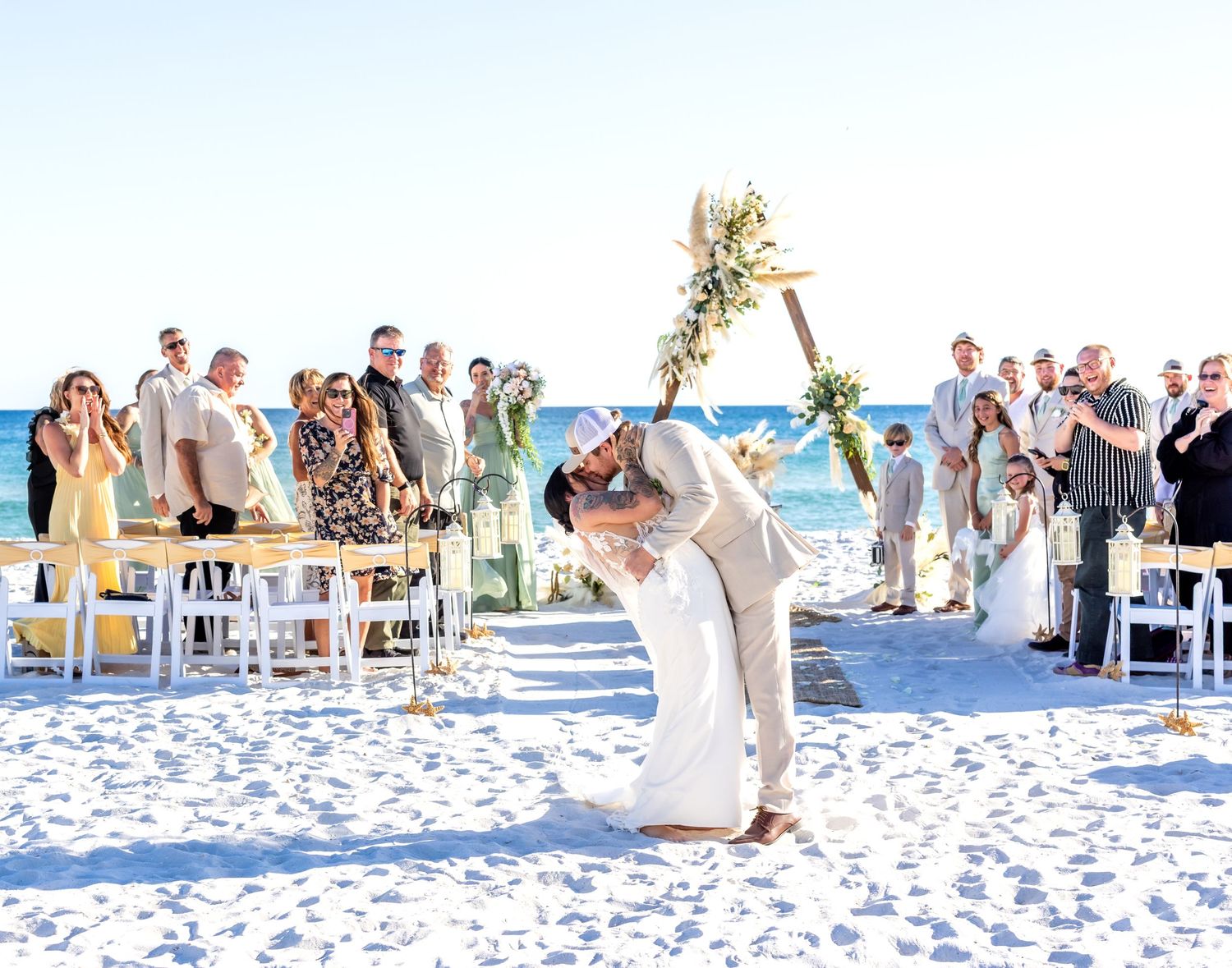 Panoramic view of a beach wedding ceremony with guests seated on white chairs along the shoreline.
