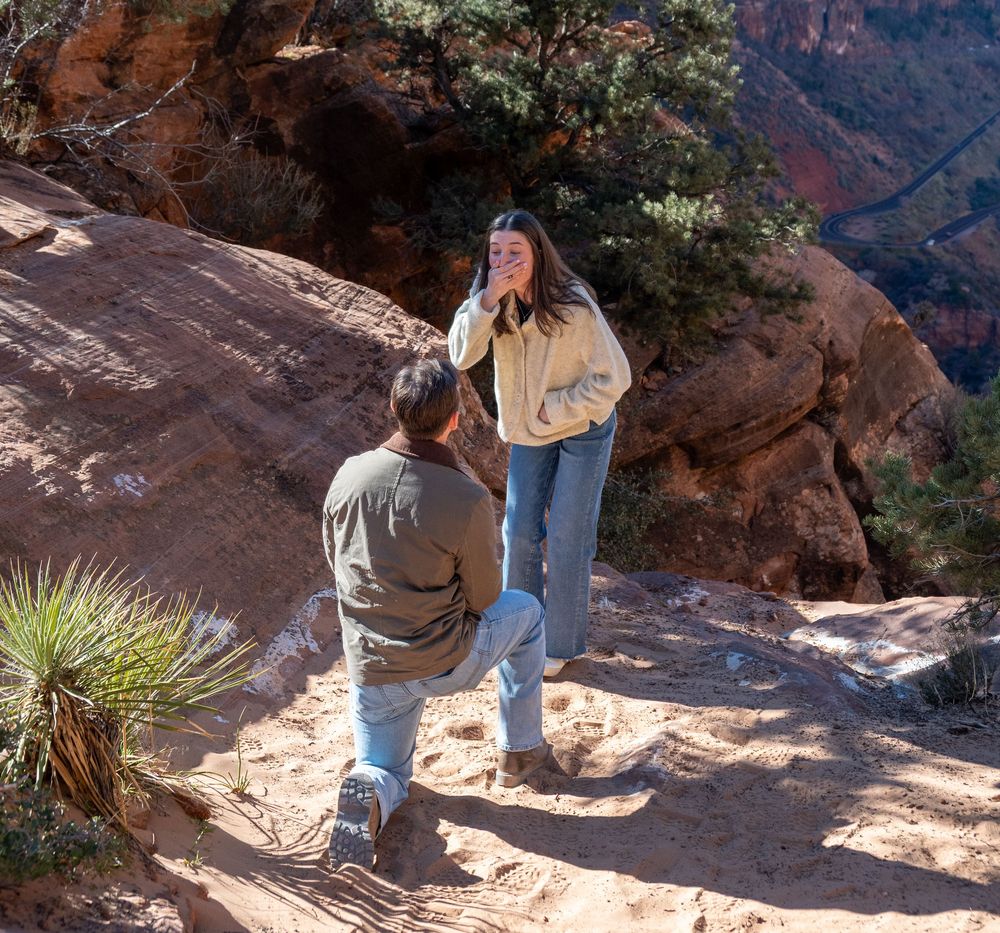 Man kneeling in marriage proposal to emotional woman on stunning Zion National Park canyon ledge.
