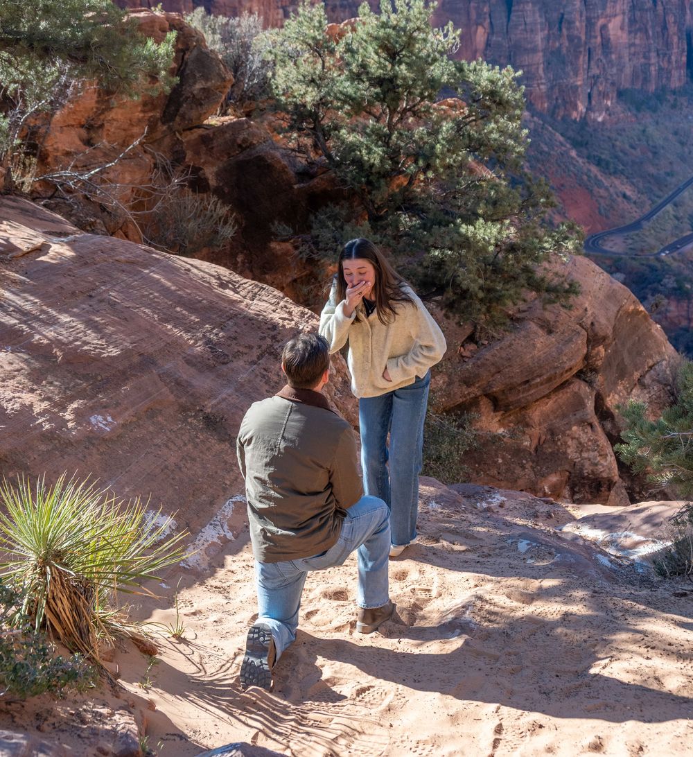 Man proposing on one knee to surprised woman on red rock trail at Zion National Park canyon overlook.