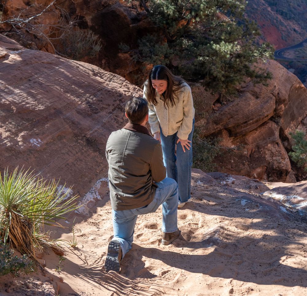 Couple during heartfelt marriage proposal on sunlit red sandstone trail at Zion National Park.