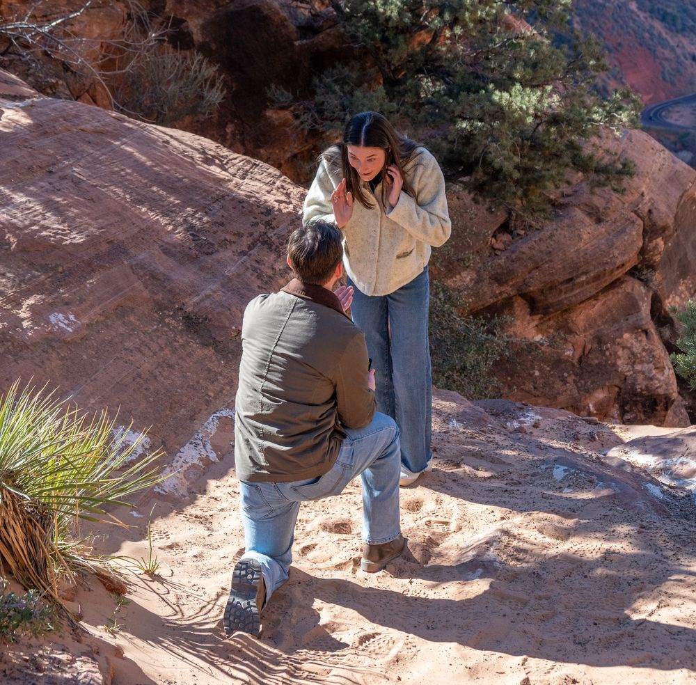 Man proposing marriage to woman on rocky desert trail with dramatic Zion canyon cliffs behind.