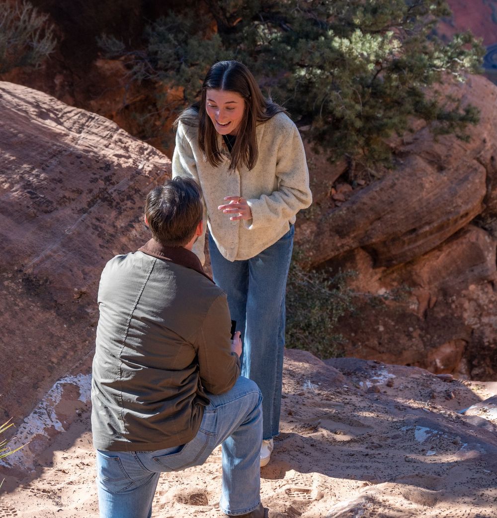 Woman smiling joyfully as partner proposes on red rock ledge at Zion National Park overlook.