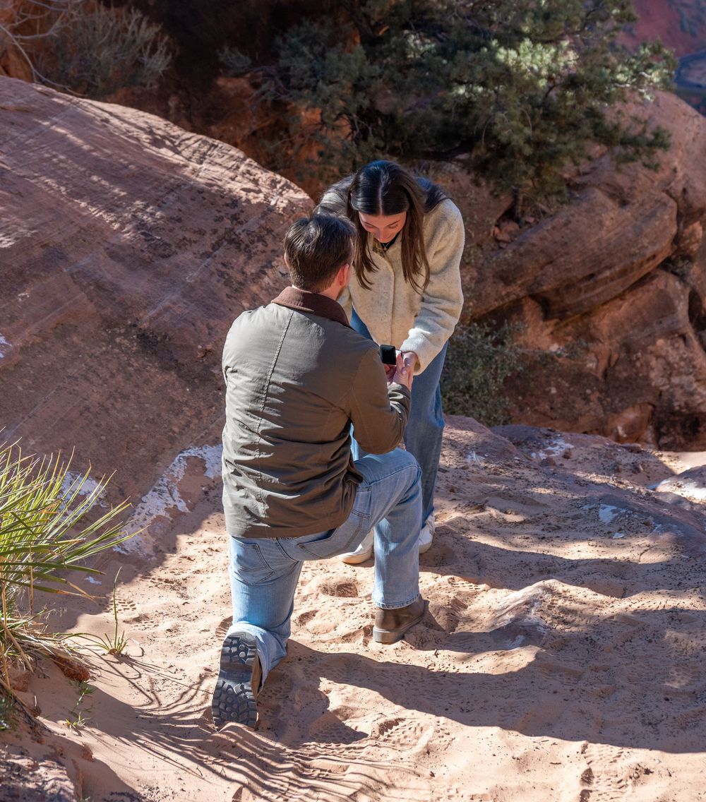 Man placing engagement ring on womans hand during proposal on Zion National Park hiking trail.