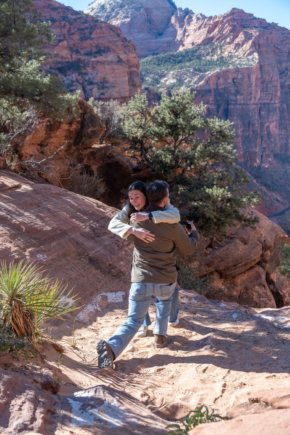 Newly engaged couple embracing happily on red sandstone trail with Zion National Park canyon views.