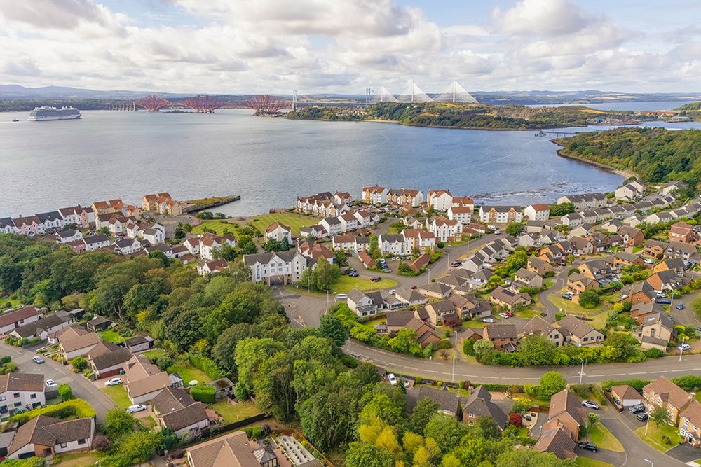 Aerial panoramic view of coastal homes and waterfront properties along a scenic bay with forth bridges in the background