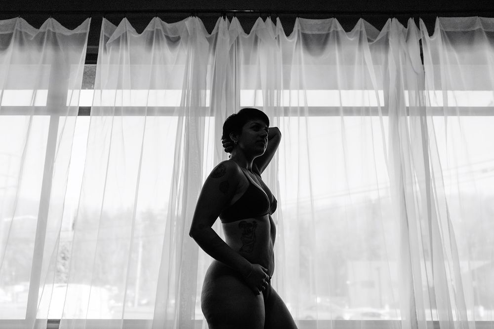 Black and white silhouette of woman in lingerie standing by sheer curtained window.