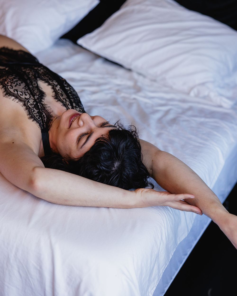 Woman with dark hair wearing black lace bra lying relaxed on white bedding.