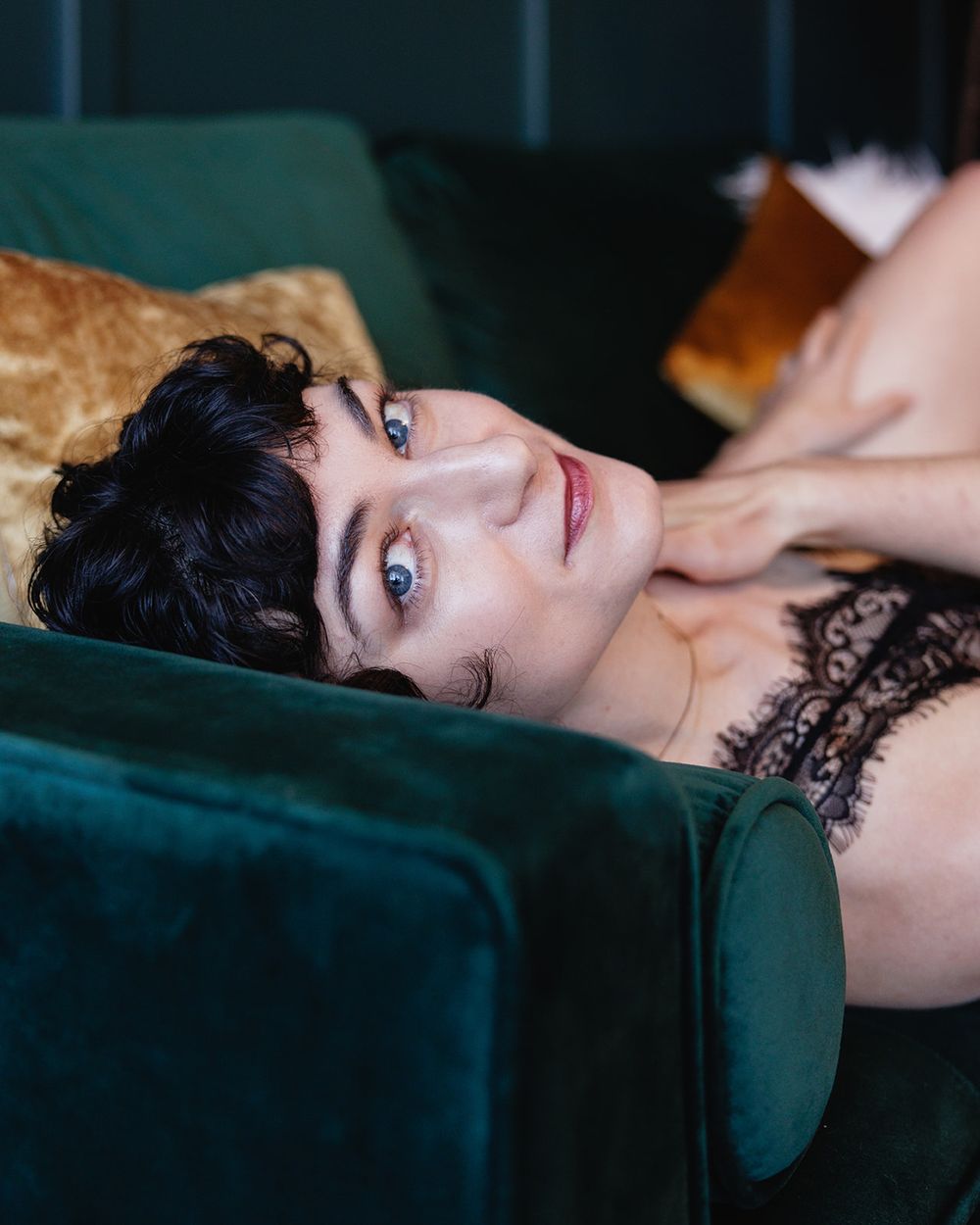 Woman with short dark curly hair and blue eyes lying on green velvet sofa in black lace.