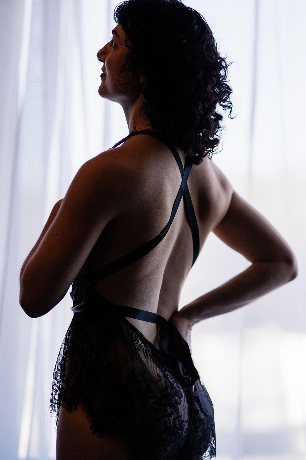 Dark silhouette of woman in open-back black lace bodysuit posing against bright window light.