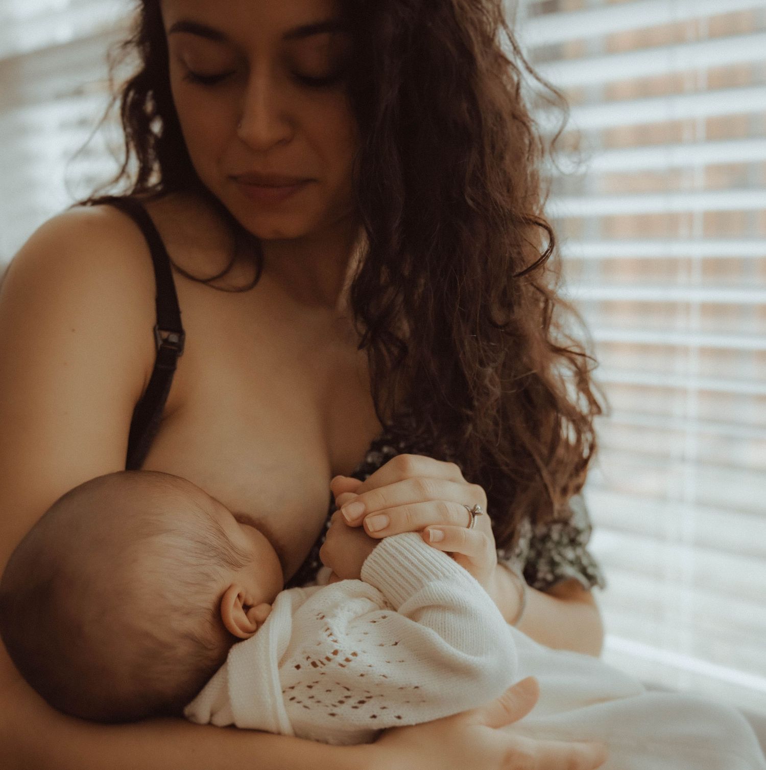 A tender moment between parent and infant in warm lighting by a window with venetian blinds.