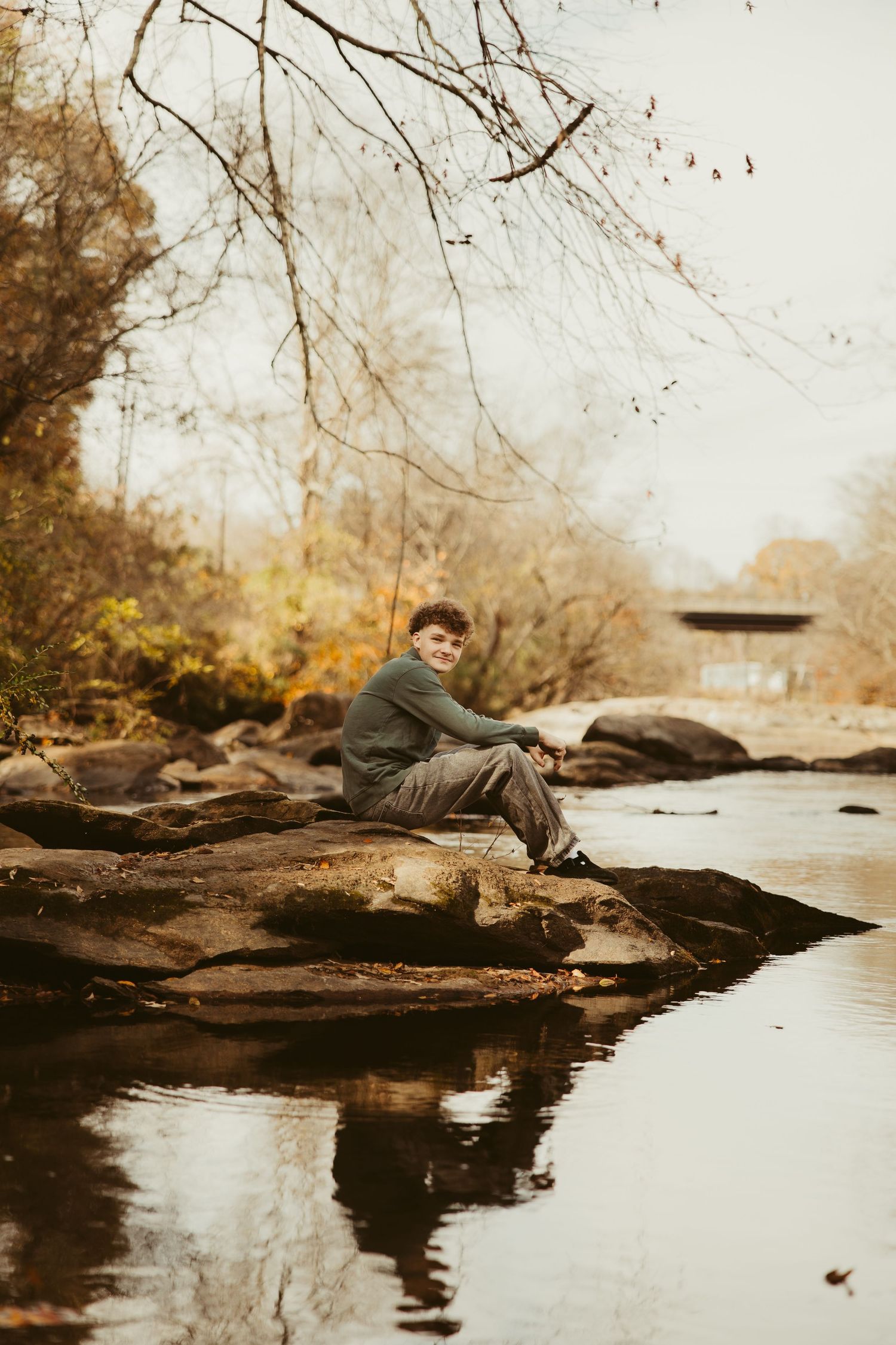 Teenage boy sitting on rocks beside a calm autumn creek with colorful fall foliage and covered bridge.