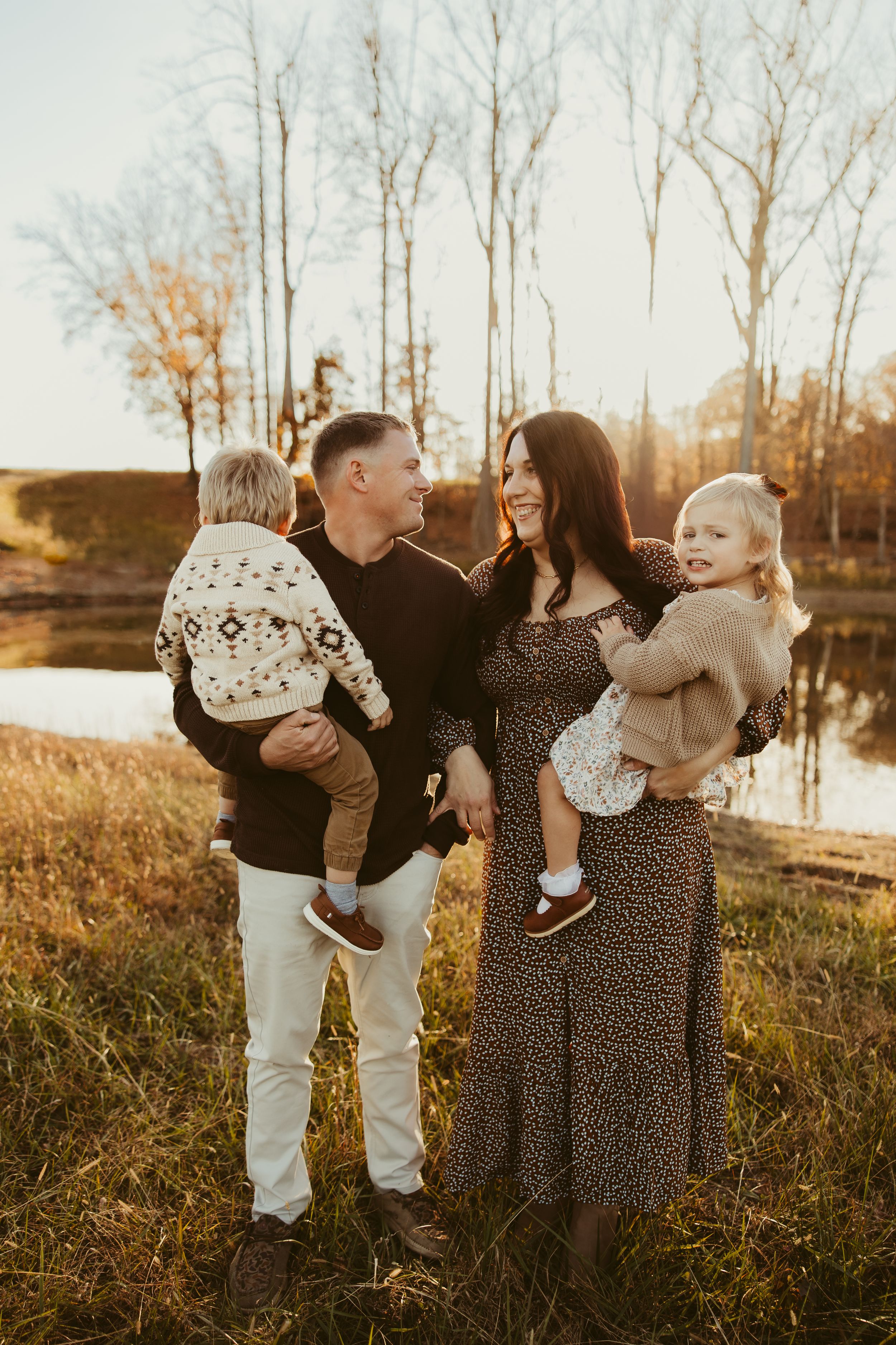 Smiling family of four with two toddlers stand by a reflective pond during warm autumn golden hour.