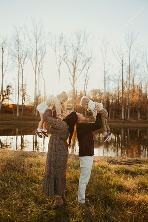 Joyful parents lift two young children in the air by a sunlit autumn pond during family photo session.