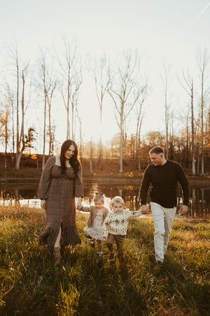 Family of four walks hand in hand through golden autumn grass near a pond at sunset.