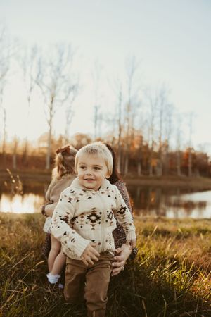 Smiling toddler boy in a patterned sweater held by a woman near an autumn lake at golden hour.