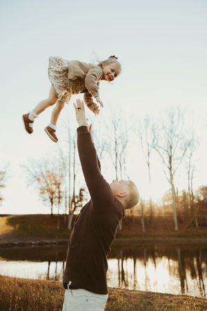 Father joyfully tossing laughing toddler girl in the air near a serene autumn lake at sunset.