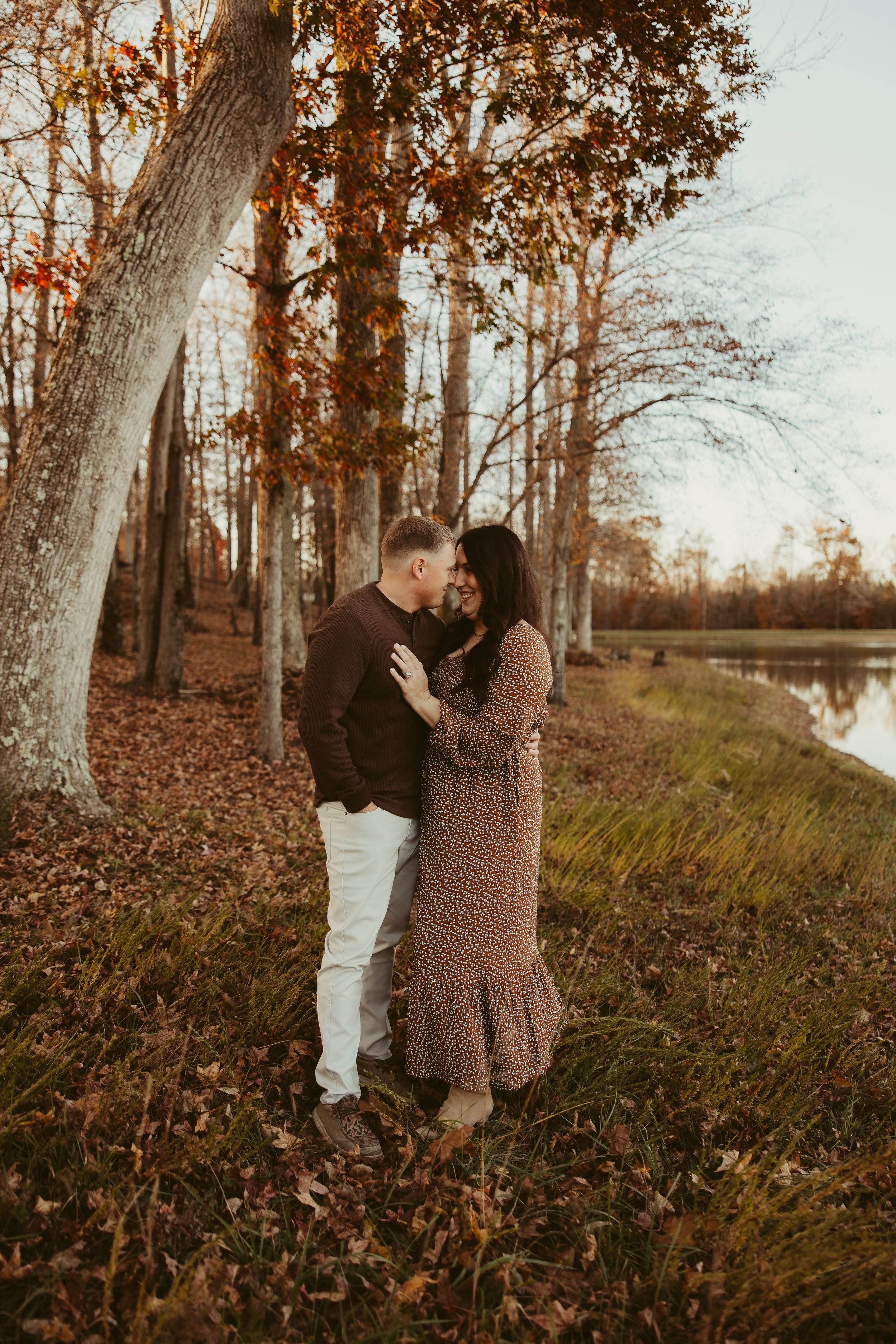 Couple sharing a tender moment embracing under autumn trees beside a peaceful lake at dusk.