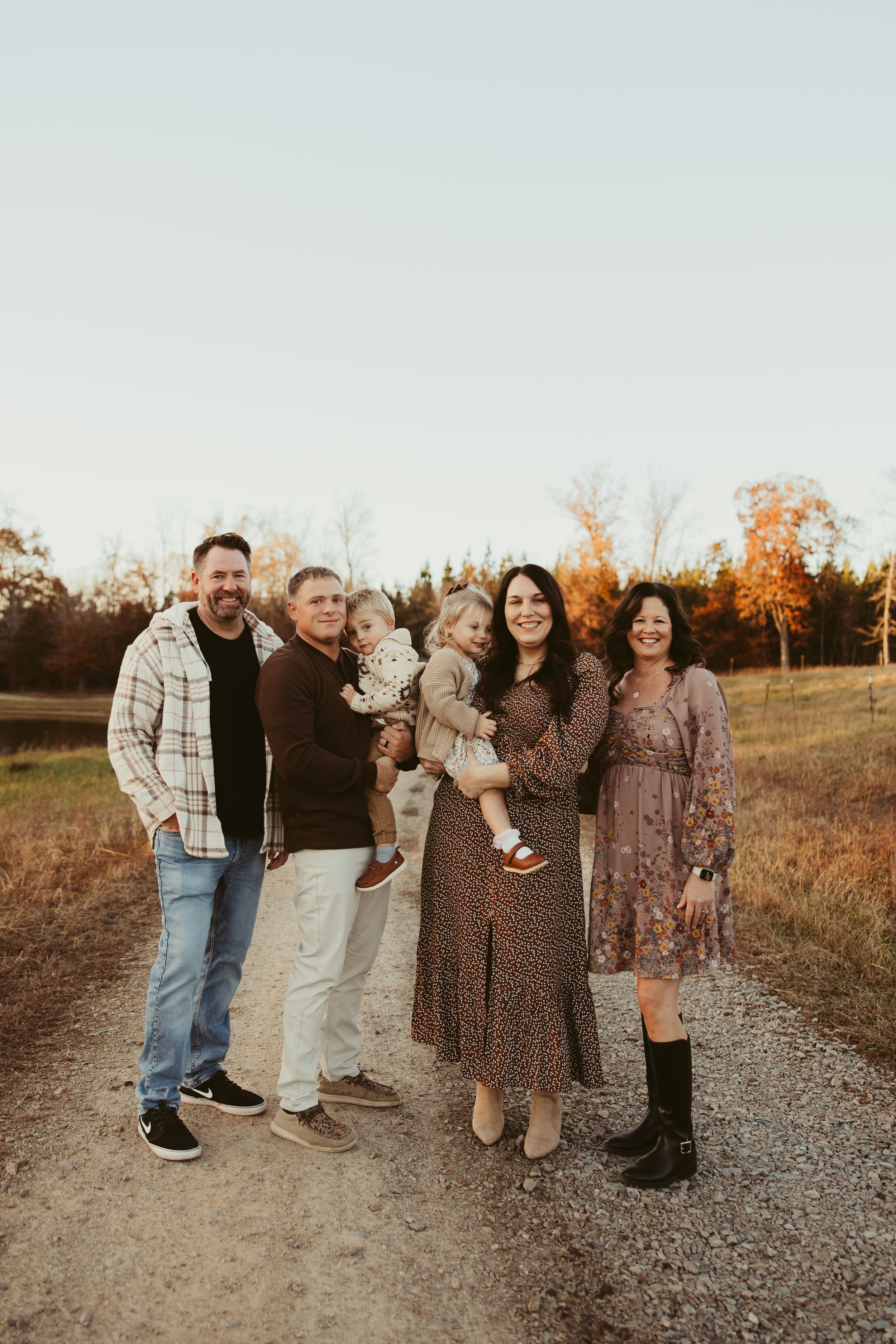 Extended family of six posing together on a gravel path with golden autumn trees in background.