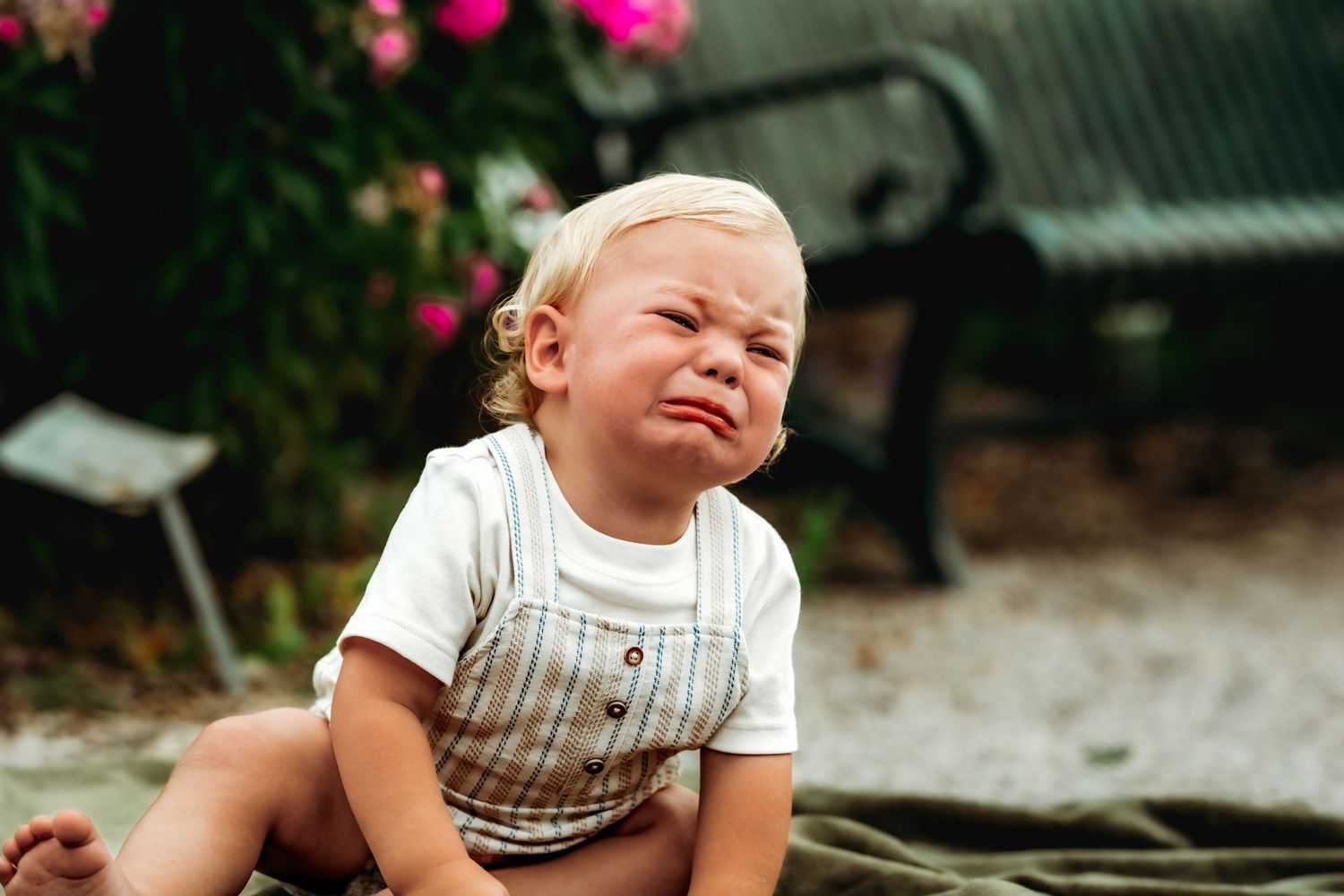 Small child in striped overalls making a funny scrunched expression while sitting outdoors near pink flowers.